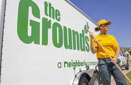 Grounds Guys employee in yellow hat and shirt, holding rake and standing next to company trailer.