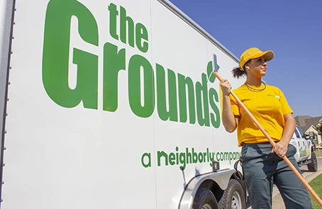 Female The Grounds Guys associate wearing branded yellow polo shirt and baseball cap standing in front of branded trailer.