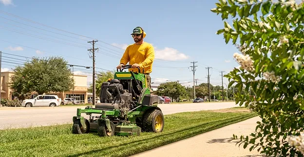 Three Grounds Guys service professionals mowing a strip of grass next to a street in a retail district.