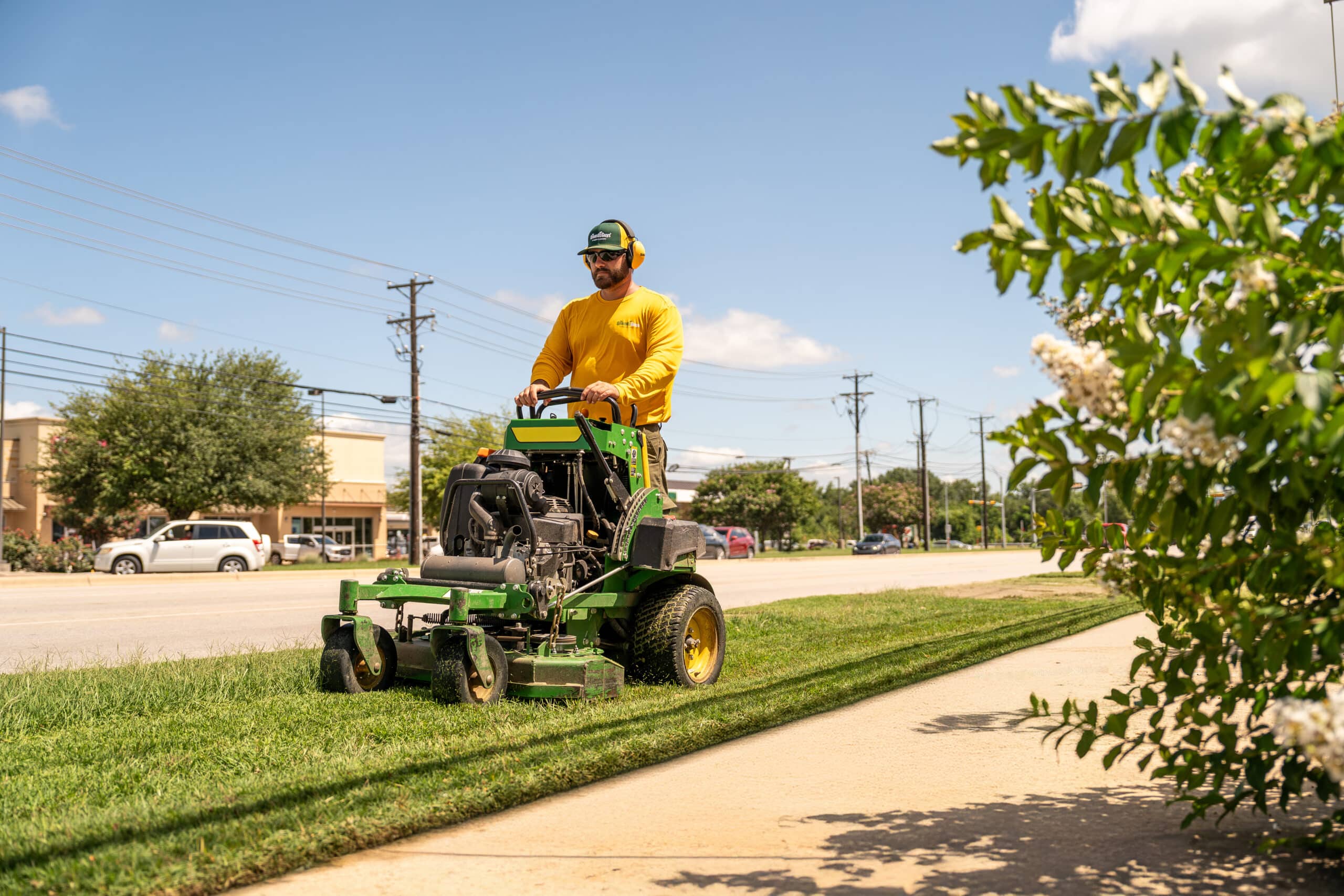 A Grounds Guys service professional operating a standing lawn mower to mow a strip of grass at a commercial property.