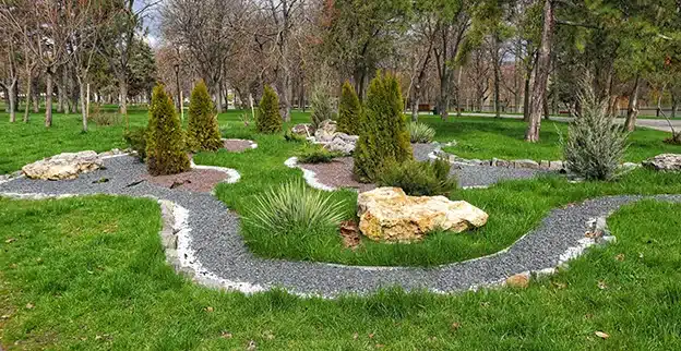 An outdoor area with gravel path, small shrubs, and decorative rock in front of a wooded area.