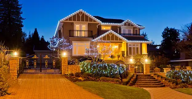 The front of a large home, with holiday lighting along its eaves and in its trees and bushes.