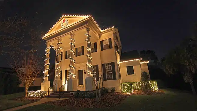 A large Colonial-style home with holiday lights and garland decorating the exterior.