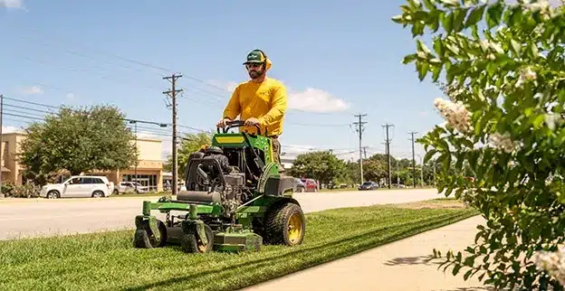 The Grounds Guys service professional mowing a strip of grass next to a street in a retail district.