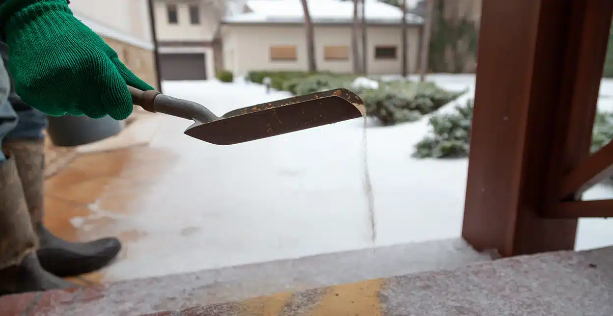 A gloved hand using a small shovel to sprinkle sand on snowy steps.