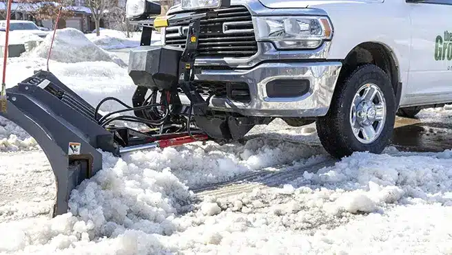 A white Grounds Guys truck, with a snow plow blade attached, pushing snow off a road.