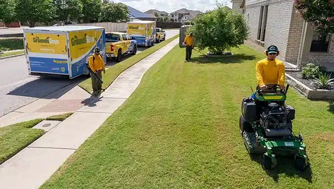 A team of The Grounds Guys service professionals cleaning up a business's lawn next to branded trucks and trailers.