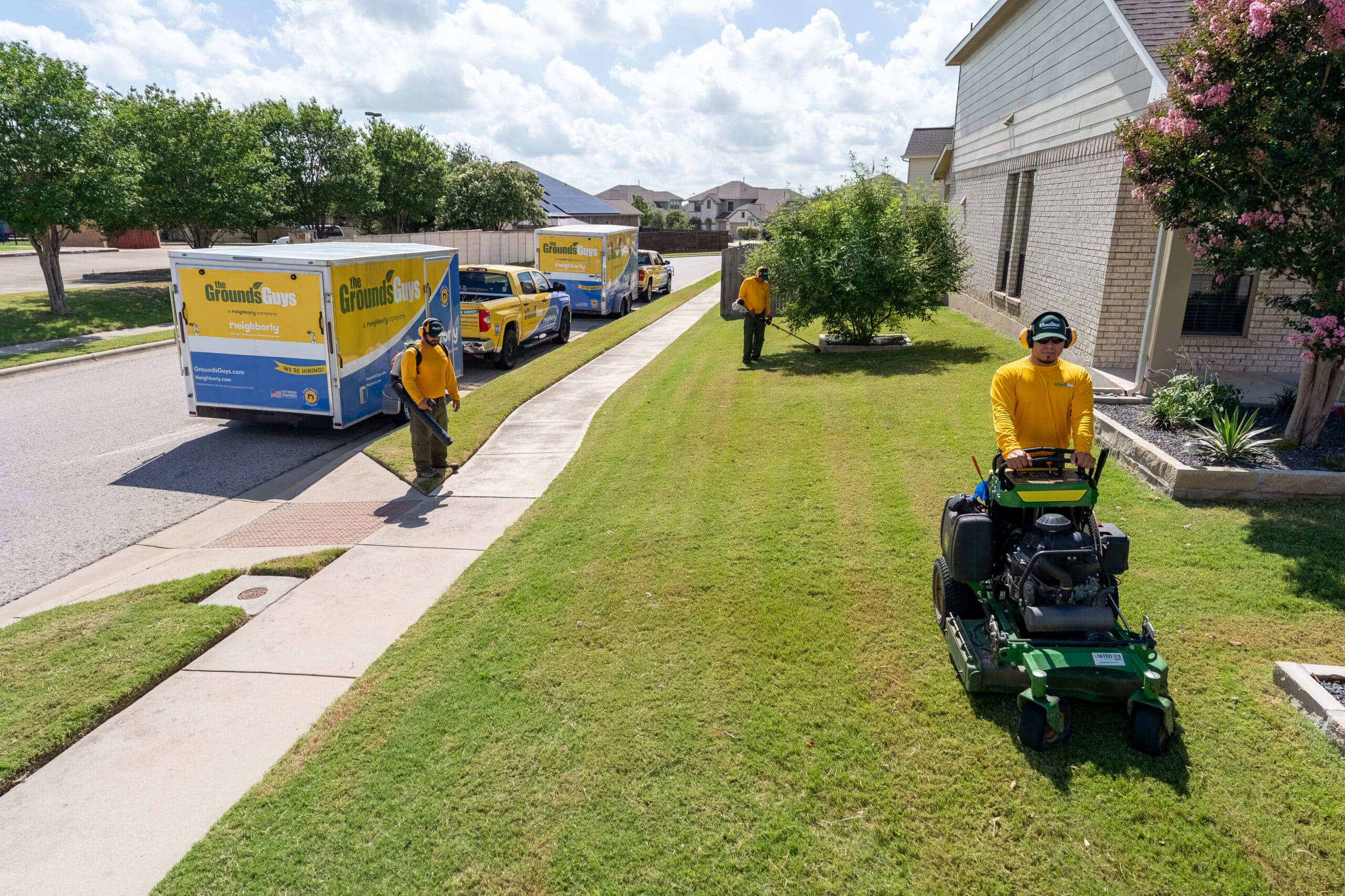 A team of The Grounds Guys service professionals mowing and edging the lawn of a home, with branded company trucks parked on the street next to the home.