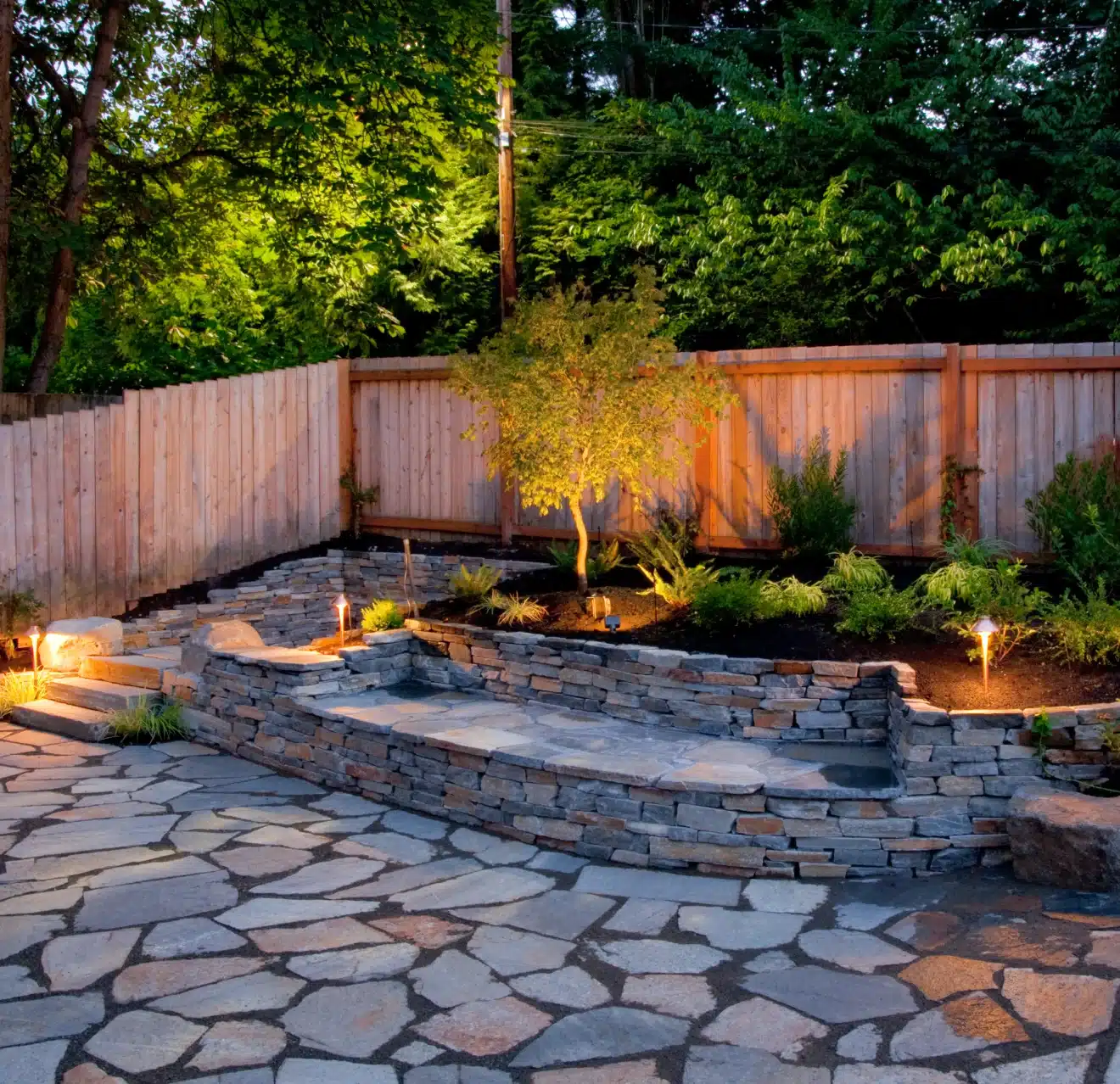 A backyard hardscape patio featuring irregular flagstone pavers and retaining wall with built-in seating, plus a small garden, wood fence, and lush trees just outside the property.