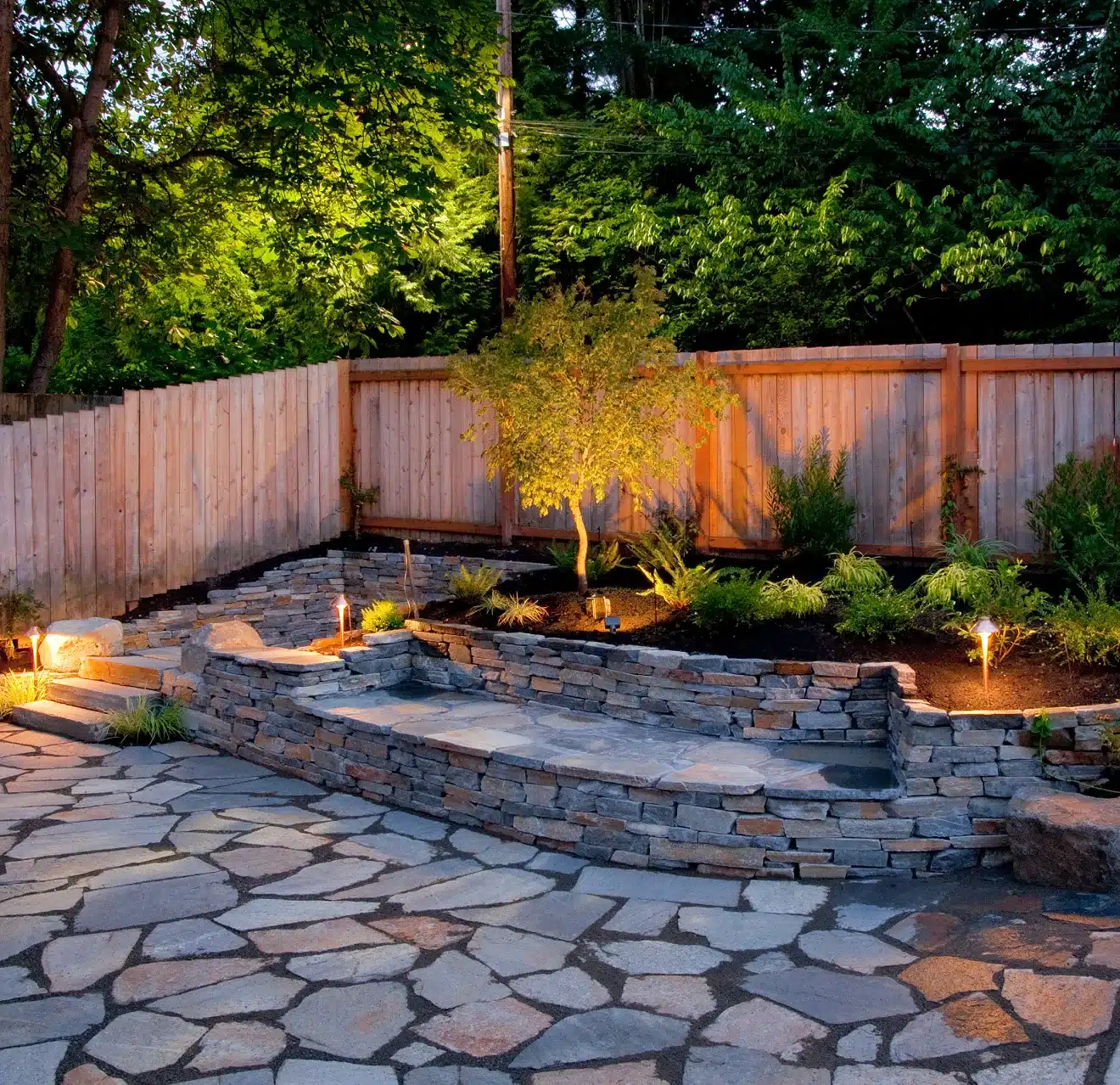 A backyard hardscape patio featuring irregular flagstone pavers and retaining wall with built-in seating, plus a small garden, wood fence, and lush trees just outside the property.