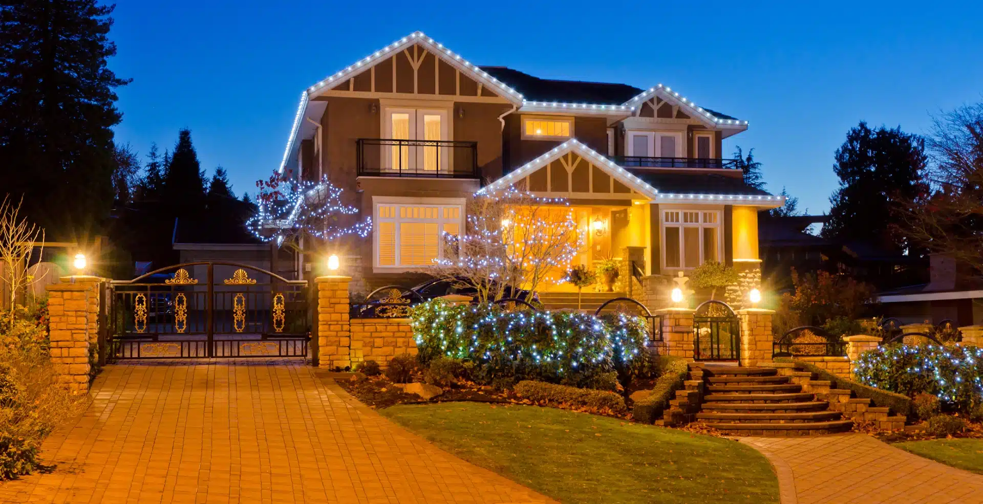 The front of a large home, with holiday lighting along its eaves and in its trees and bushes.