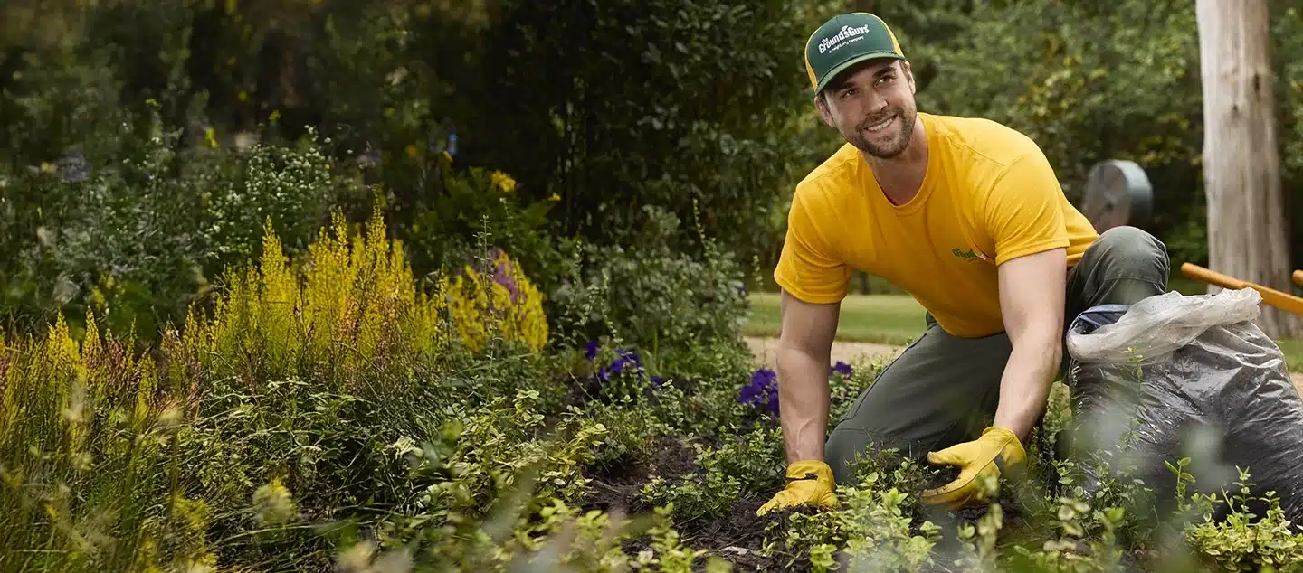 A Grounds Guys service professional smiling while kneeling in a garden bed to tend to the wide variety of plants.