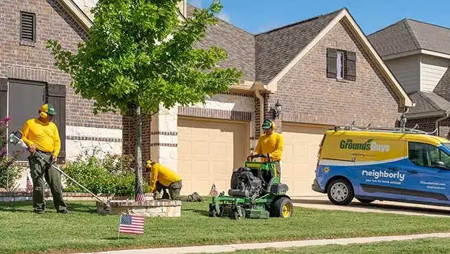 Two Grounds Guys service professionals mowing and edging a home's front lawn, with a branded van parked in the driveway.
