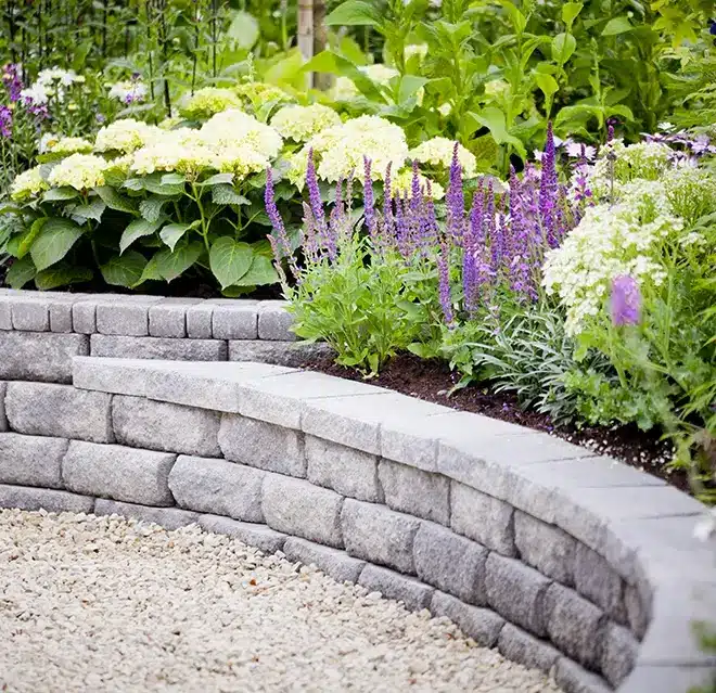 A plant bed with a variety of shrubs and plants accented by column lights and framed by stone blocks.