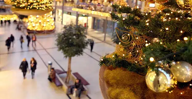 Holiday lighting and decorations hung from the upper story of a mall, with shoppers visible below.