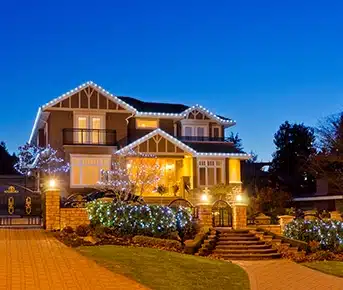 The front of a large home, with holiday lighting along its eaves and in its trees and bushes.