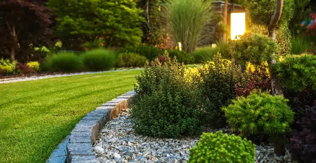 A well-landscaped rock garden bed along the border of a well-maintained lawn, with small shrubs, plants, and decorative lighting.