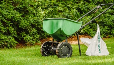 Manual walk behind grass seed spreader and bag of lawn fertilizer sitting on a green lawn in a residential home.