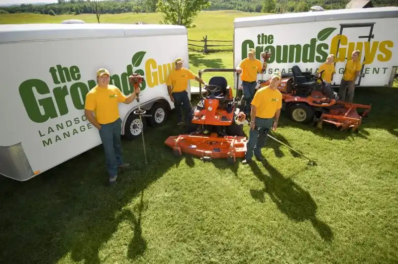 Grounds Guys crew posing with riding lawnmowers and branded trailers.