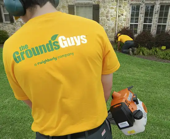 Grounds Guys employee wearing t-shirt with logo on back and using leaf blower to clean up client's front yard. 