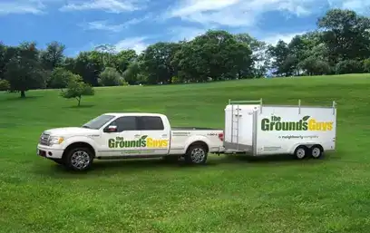 Branded Grounds Guys truck and trailer parked in grassy field.