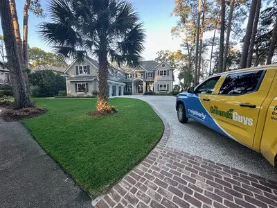 Large home with bright green front yard and Grounds Guys vehicle parked in wrap-around driveway.
