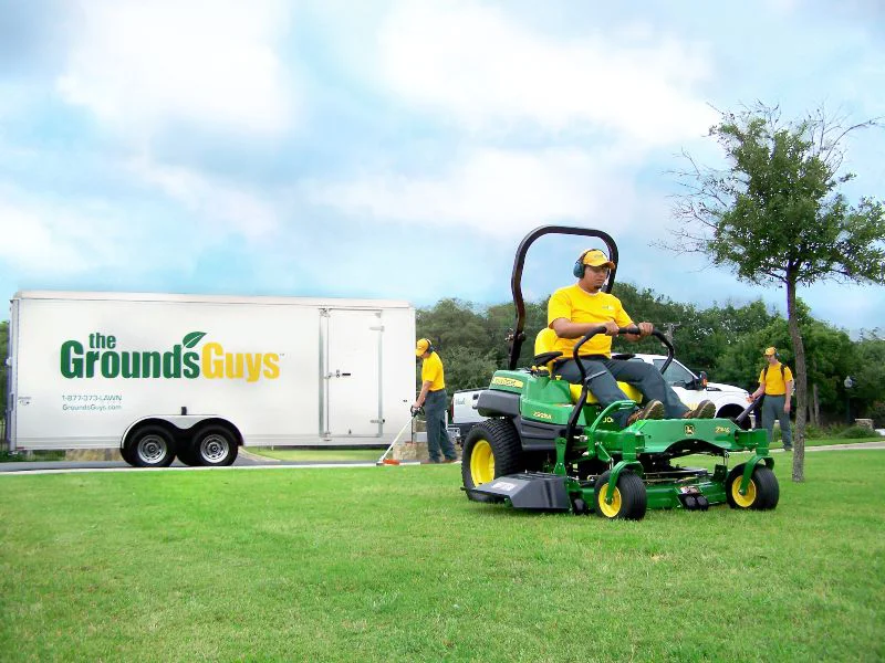 The Grounds Guys service professional on riding lawn mower with branded trailer behind him.
