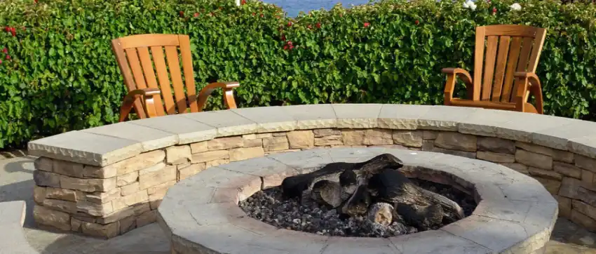Stone firepit with wraparound stone bench, Adirondack chairs, and hedge.