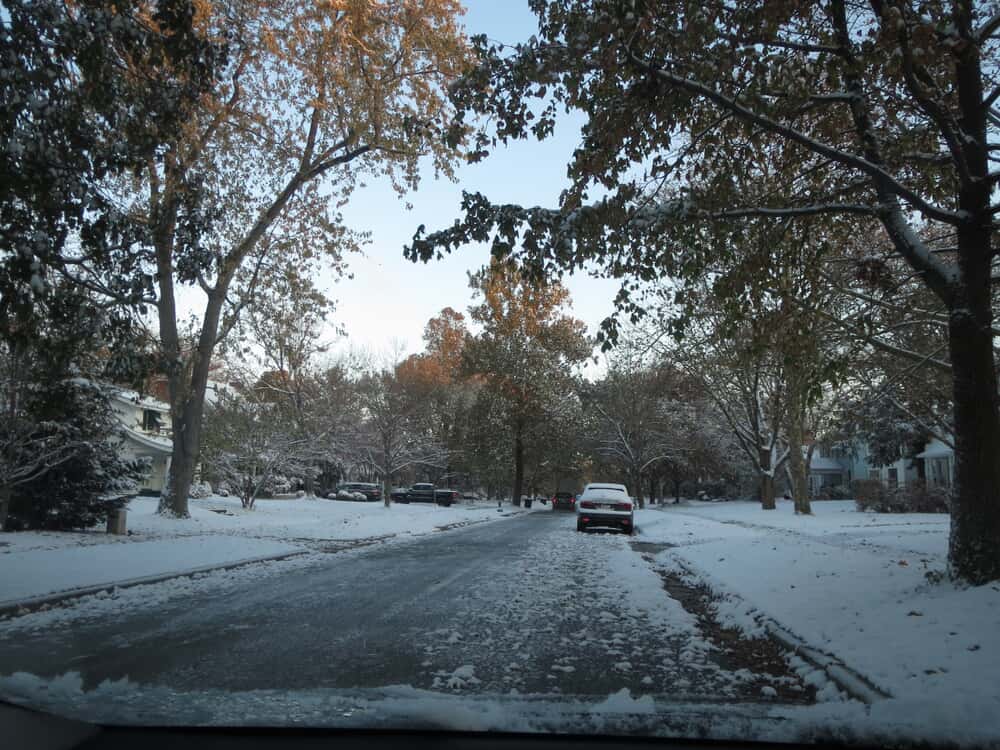 Snow on an empty residential street with a car and trees.