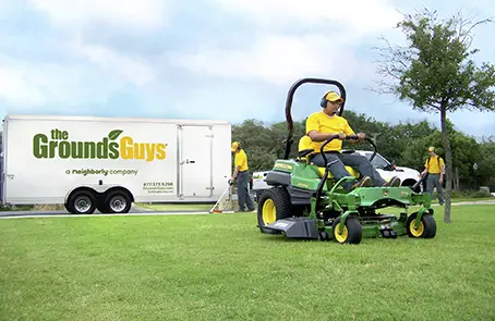 Grounds Guys associate riding lawn mower with branded trailer in background.